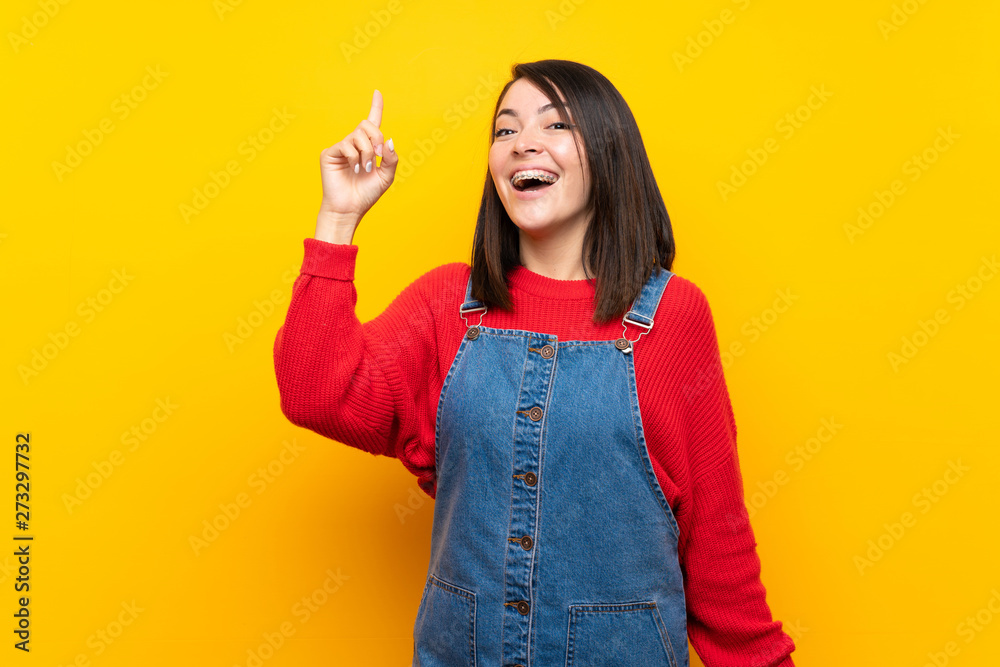 Young Mexican woman with overalls over yellow wall intending to realizes the solution while lifting a finger up Young Mexican woman with overalls over yellow wall intending to realizes the solution while lifting a finger up