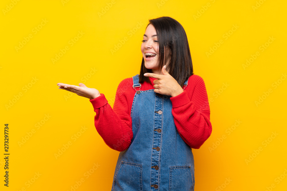 Young Mexican woman with overalls over yellow wall holding copyspace imaginary on the palm to insert an ad Young Mexican woman with overalls over yellow wall holding copyspace imaginary on the palm to insert an ad