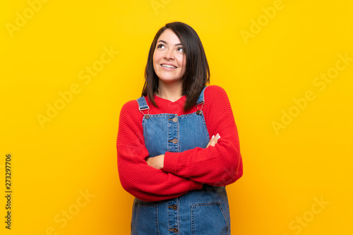 Young Mexican woman with overalls over yellow wall looking up while smiling © luismolinero