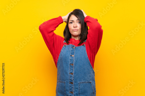 Young Mexican woman with overalls over yellow wall frustrated and takes hands on head © luismolinero