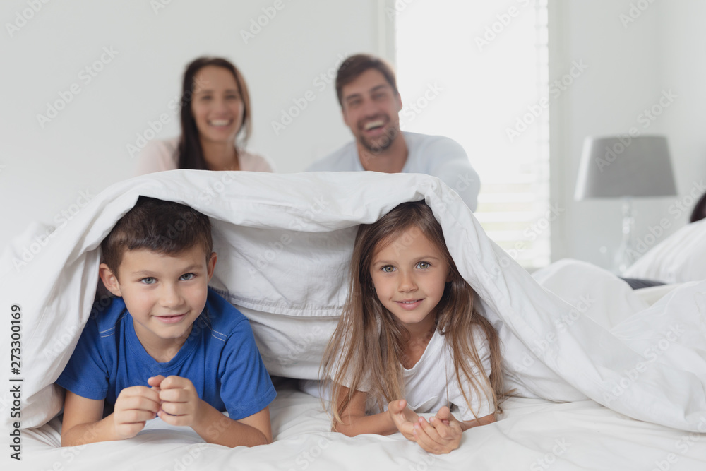 Children lying under blanket while parent sitting on bed in bedroom at home