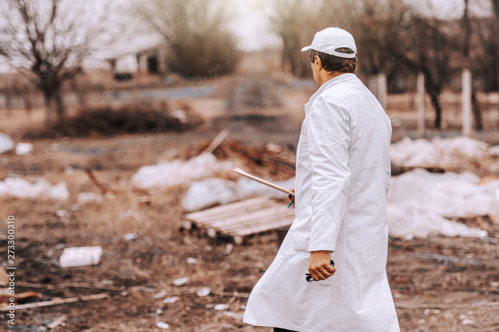 Ecologist in white uniform and cap on head holding clipboard while walking on landfill. Backs turned. Ecologist in white uniform and cap on head holding clipboard while walking on landfill. Backs turned.
