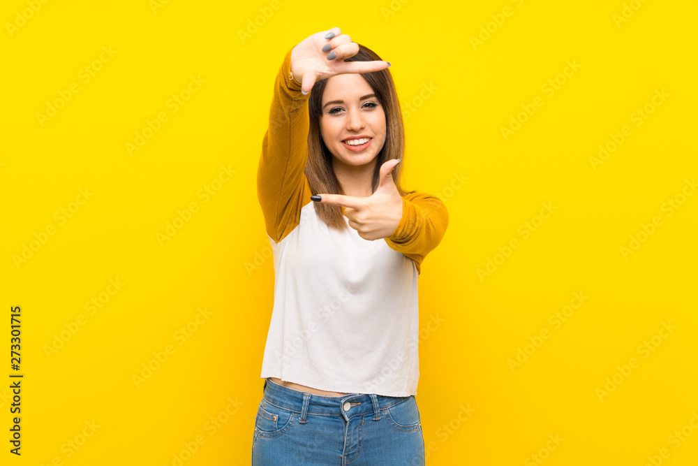 Pretty young woman over isolated yellow wall focusing face. Framing symbol Pretty young woman over isolated yellow wall focusing face. Framing symbol