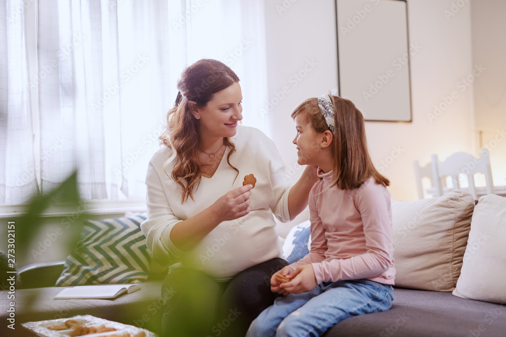 Beautiful happy mother eating cookies with her loving daughter in living room.
