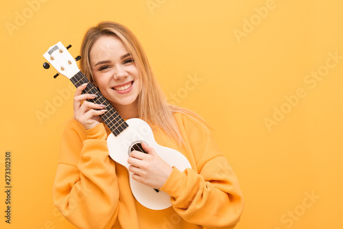 Smiling blonde in yellow clothes with ukulele in hand is isolated on an orange background, looking into the camera and rejoicing. Portrait of a positive girl with Hawaiian guitar in hand. Copy space