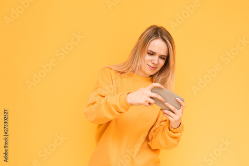 Concentrated girl in yellow clothes playing video games on a smartphone, focused on looking at the screen. Blonde gamer plays mobile games on a yellow background. Copy space
