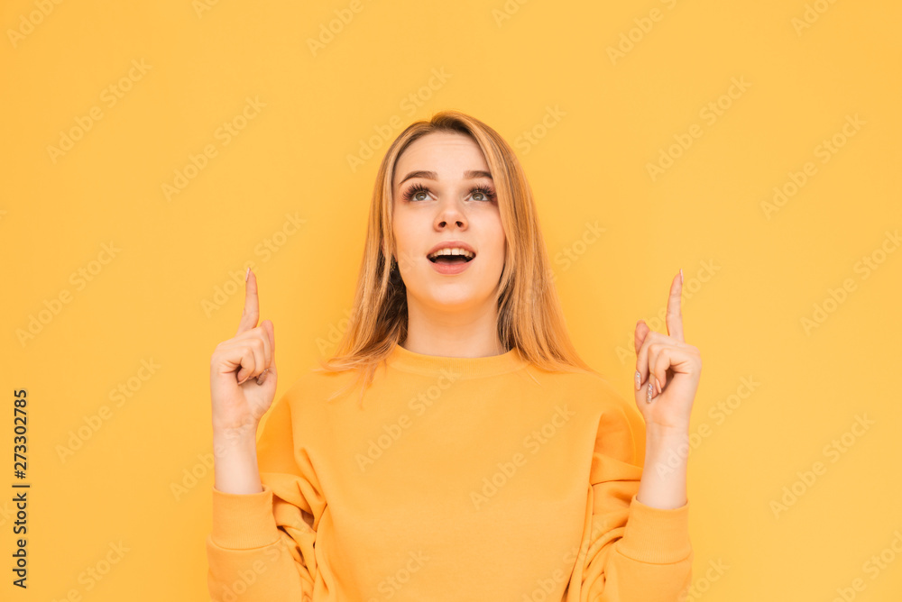 Emotional girl in a bright casual clothing looks and shows fingers up on a blank space, shocking face, isolated on a yellow background. Surprised teenage girl shows fingers up on copyspace Emotional girl in a bright casual clothing looks and shows fingers up on a blank space, shocking face, isolated on a yellow background. Surprised teenage girl shows fingers up on copyspace