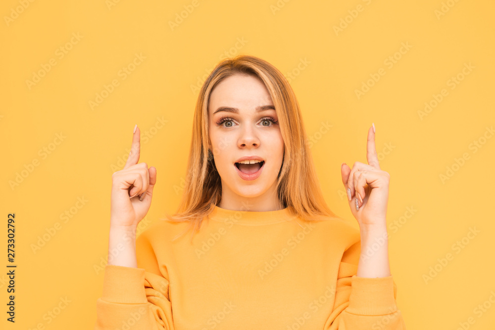 Attractive blonde with a shocking face is isolated on a yellow background, looks at the camera with astonishment and shows fingers up. Surprised girl shows her fingers in an empty place. Copy space