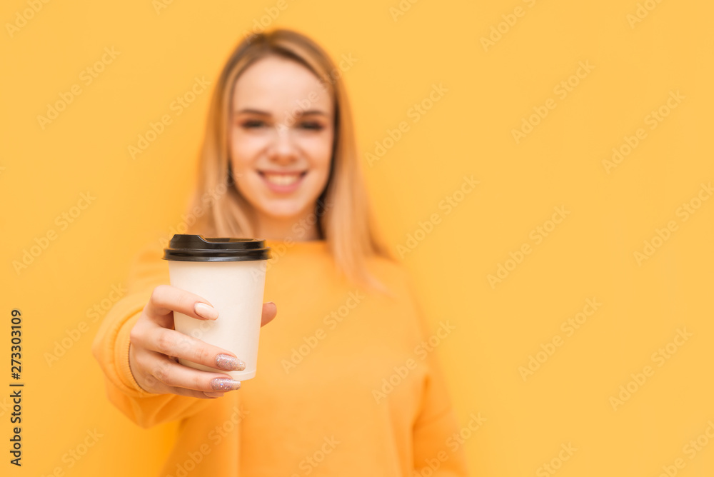 Happy girl holds a paper glass of coffee in her hand and shows her in the camera on a yellow background, smiling and looking. Focus on a cup of coffee in the hands of a young girl underground. Happy girl holds a paper glass of coffee in her hand and shows her in the camera on a yellow background, smiling and looking. Focus on a cup of coffee in the hands of a young girl underground.