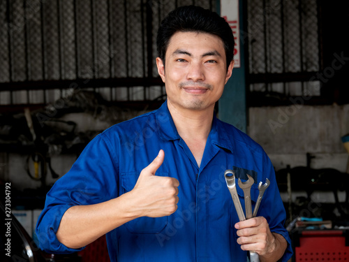 Young asian auto mechanic in uniform holding wrenches and showing thumbs up in the garage. Repair, car service and maintenance concept.