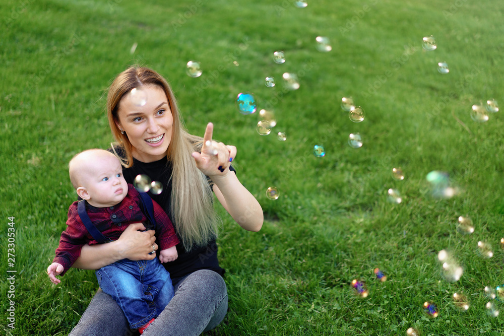 Young Caucasian mother inflates soap bubbles with her little son in a park on a sunny summer day. Young Caucasian mother inflates soap bubbles with her little son in a park on a sunny summer day.