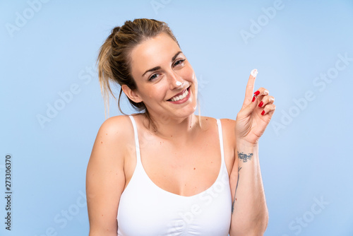 Happy Young blonde woman with moisturizer over isolated blue wall