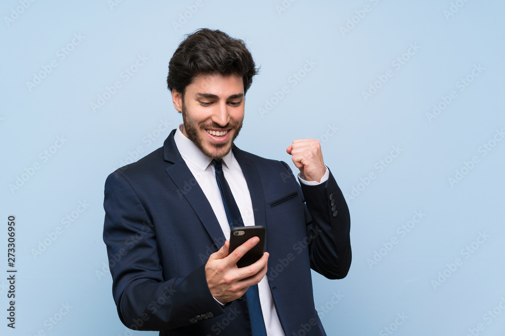 Businessman over isolated blue wall with phone in victory position Businessman over isolated blue wall with phone in victory position