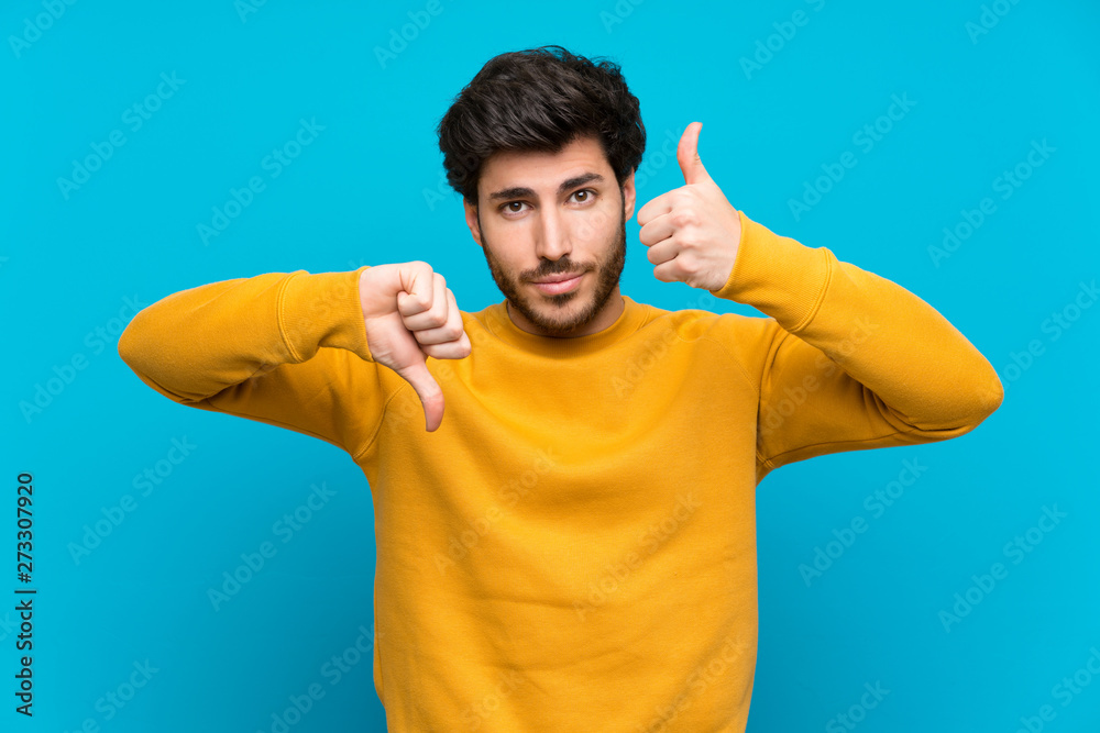 Handsome over isolated blue wall making good-bad sign. Undecided between yes or not Handsome over isolated blue wall making good-bad sign. Undecided between yes or not
