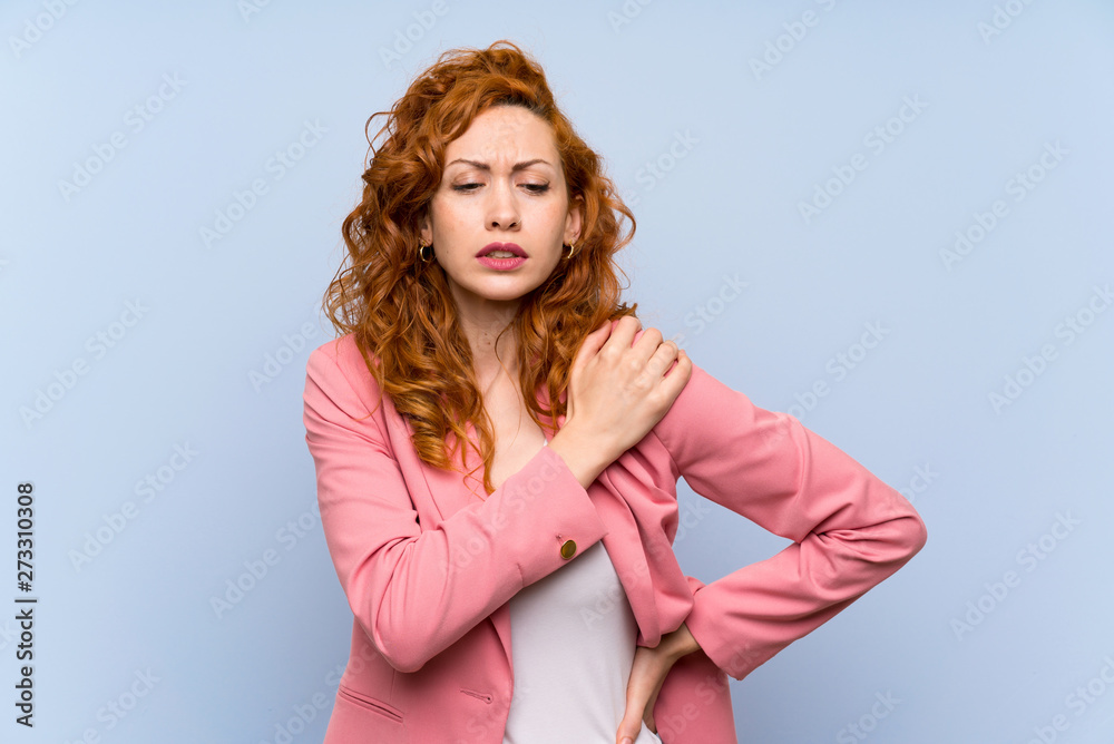 Redhead woman in suit over isolated blue wall suffering from pain in shoulder for having made an effort Redhead woman in suit over isolated blue wall suffering from pain in shoulder for having made an effort