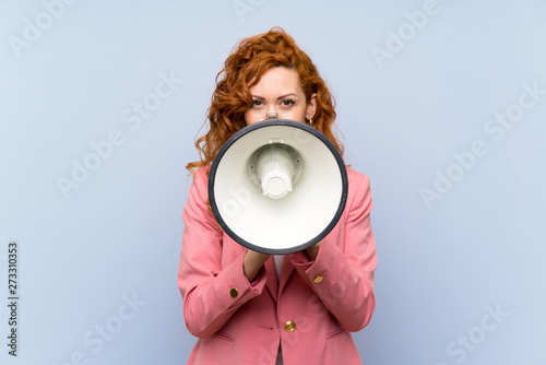 Redhead woman in suit over isolated blue wall shouting through a megaphone