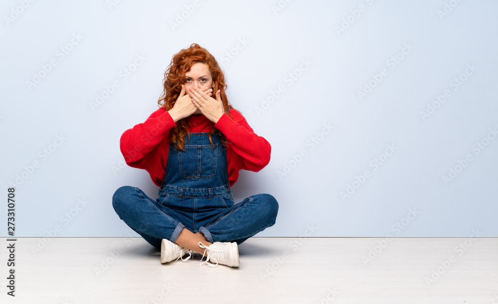 Redhead woman with overalls sitting on the floor covering mouth with hands