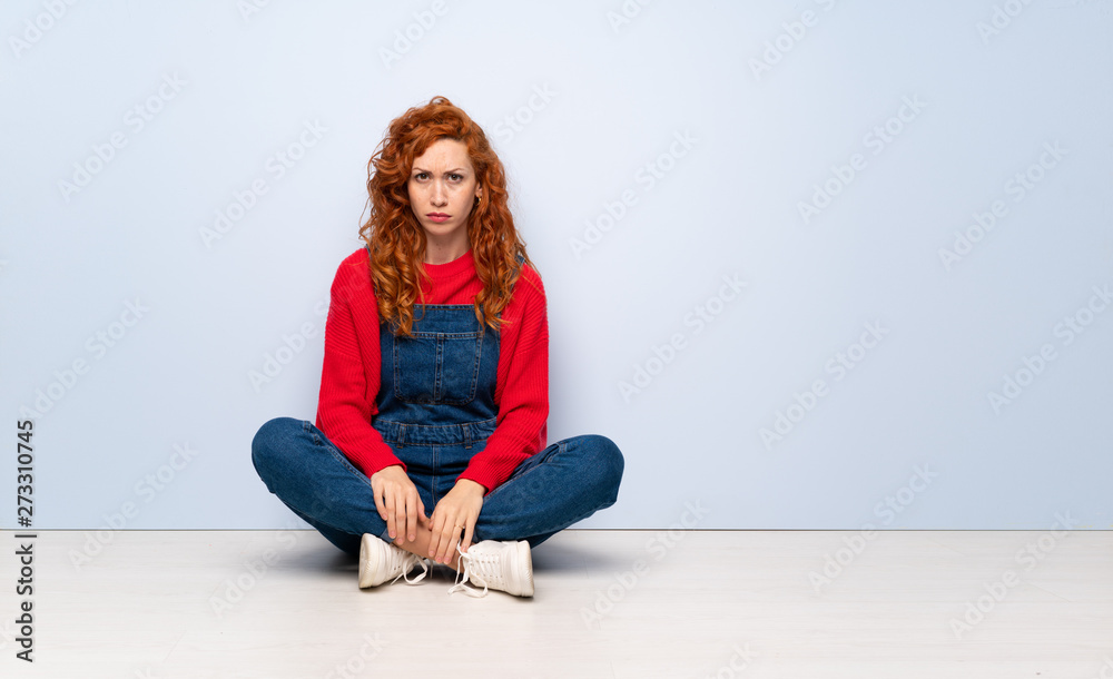 Redhead woman with overalls sitting on the floor with sad and depressed expression Redhead woman with overalls sitting on the floor with sad and depressed expression