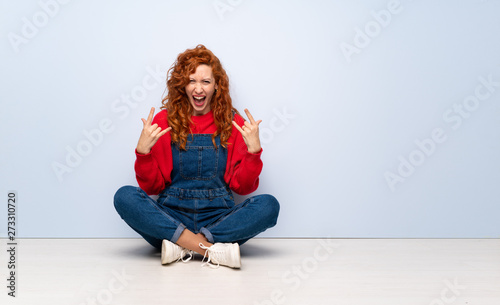 Redhead woman with overalls sitting on the floor making rock gesture