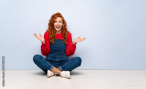 Redhead woman with overalls sitting on the floor with shocked facial expression