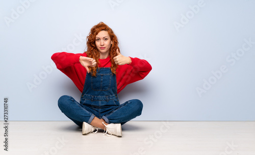 Redhead woman with overalls sitting on the floor making good-bad sign. Undecided between yes or not