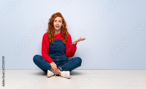 Redhead woman with overalls sitting on the floor making doubts gesture