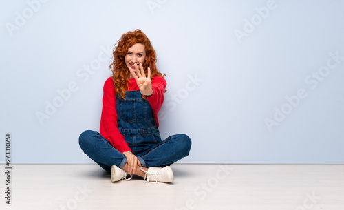 Redhead woman with overalls sitting on the floor happy and counting four with fingers