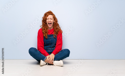 Redhead woman with overalls sitting on the floor shouting to the front with mouth wide open