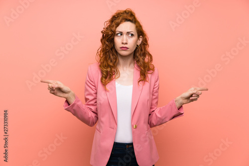 Fotografia Redhead woman in suit over isolated pink wall pointing to the laterals having do