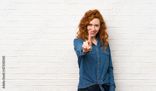 Redhead woman over white brick wall showing and lifting a finger