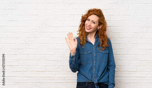 Redhead woman over white brick wall saluting with hand with happy expression