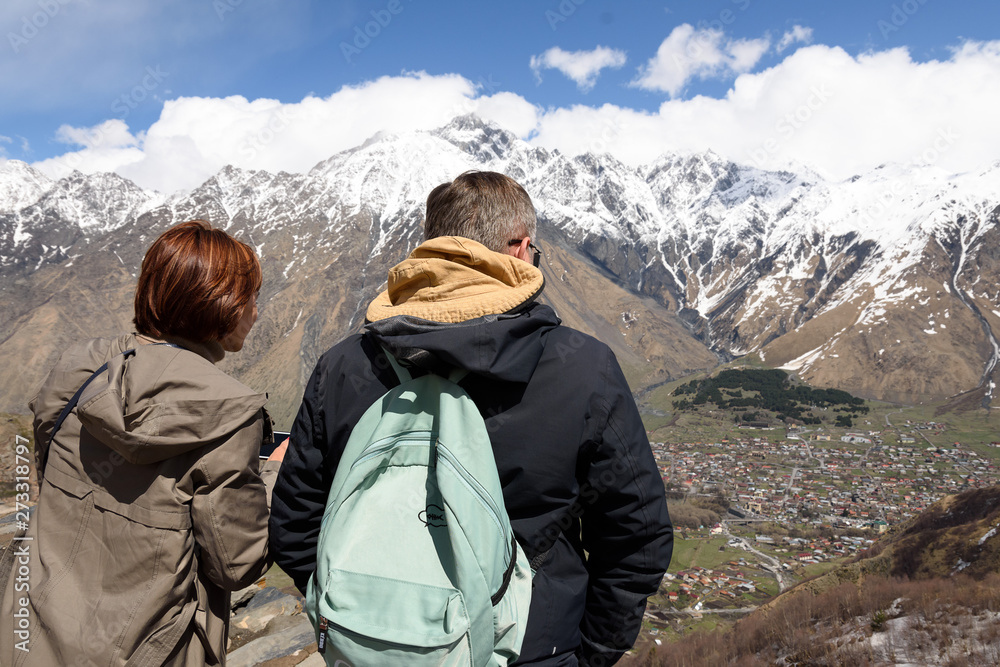 Georgian Military Road, GE-May,3 2019: Two tourists on mountain over Stepantsminda township, highland region of Georgia, main Caucasian ridge. Second name of village - Kazbegi