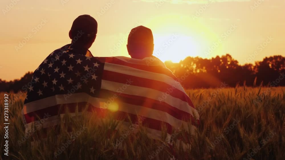 A man and his son admire the sunset over a field of wheat, wrapped in the flag of the USA