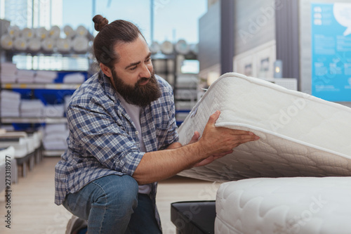 Household shopping concept. Mature bearded man examining orthopedic mattress at furnishings store. Handsome male customer choosing new bed at furniture shop