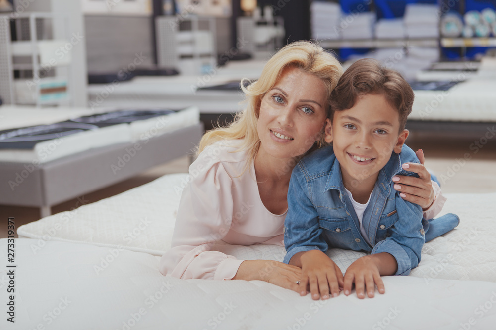 Gorgeous happy mature woman embracing her lovely son, smiling to the camera, trying new comfortable orthopedic bed at furniture store. Mother and son shopping for furniture for their home Gorgeous happy mature woman embracing her lovely son, smiling to the camera, trying new comfortable orthopedic bed at furniture store. Mother and son shopping for furniture for their home