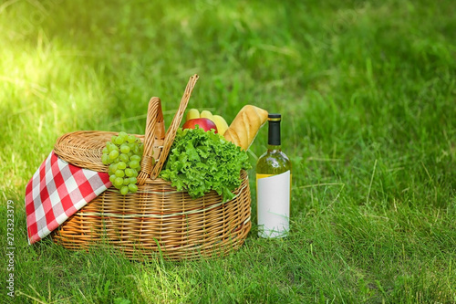 Wicker basket with blanket  wine and food on green grass in park  space for text. Summer picnic