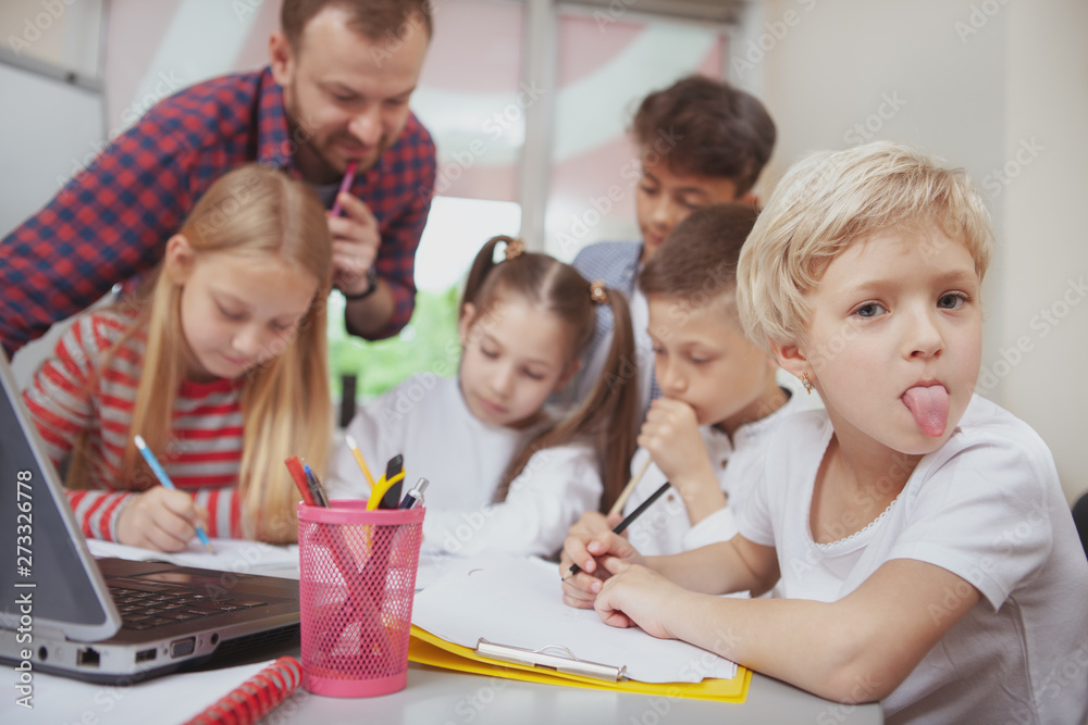 Cute little blond haired girl sticking her tongue out, having fun at school, her classmates and teacher talking on the background Cute little blond haired girl sticking her tongue out, having fun at school, her classmates and teacher talking on the background