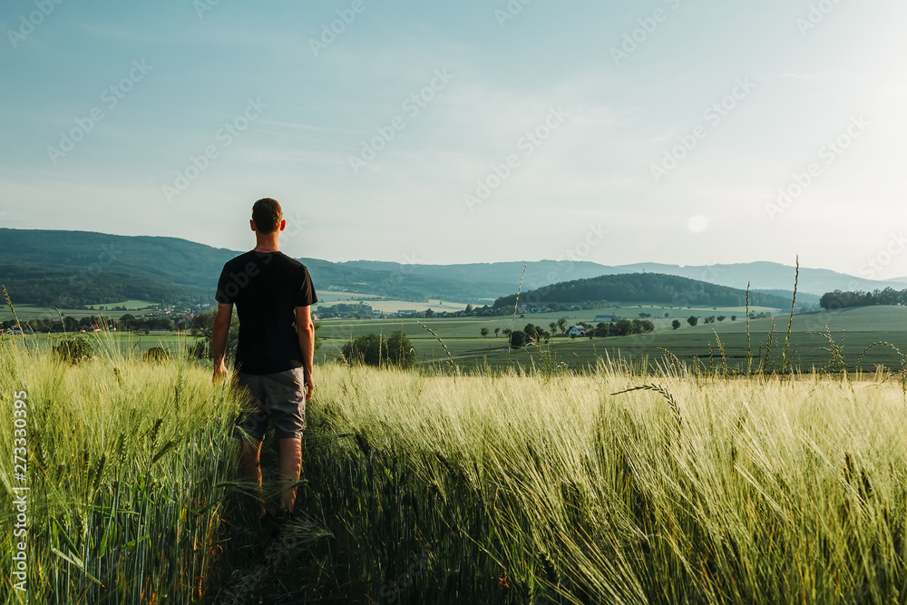 Young man standing in rural czech landscape with wheat field, hill and trees at sunset