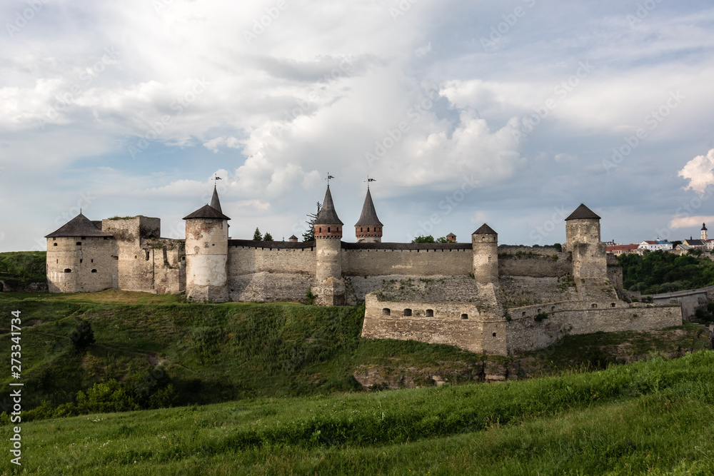 A view on the side of the medieval Kamianets-Podilskyi fortress of the XVI century, located on a rocky green hill, under a beautiful cloudy sky. Ukraine.