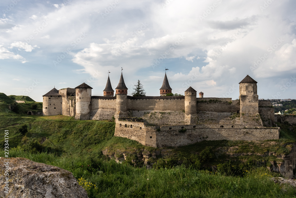 A side view of the medieval Kamianets-Podilskyi fortress of the XVI century, located on a rocky green hill, under the cloudy sky. On the foreground a stone is visible. Ukraine.