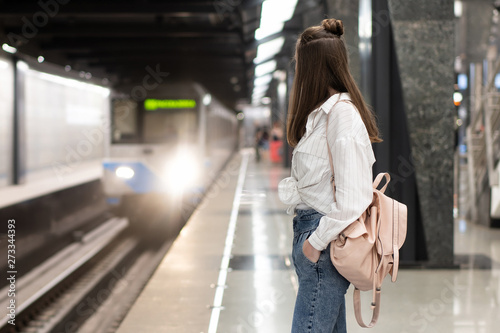 young european girl in jeans and shirt with backpack is waiting for the train at the metro station. Background in motion blur to convey a vibrant atmosphere. This is a snapshot idea.