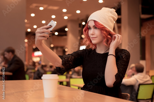 Beautiful girl with orange hair, in a cap photographes herself on a smartphone. Selfie on the phone. Sitting to wait friends alone at a food court in a cafe. Fashionable image. People Z. photo