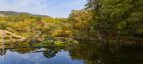 Japanese garden, a cottage hidden among japanese maple trees and pines. A small island on a pond in foreground.
