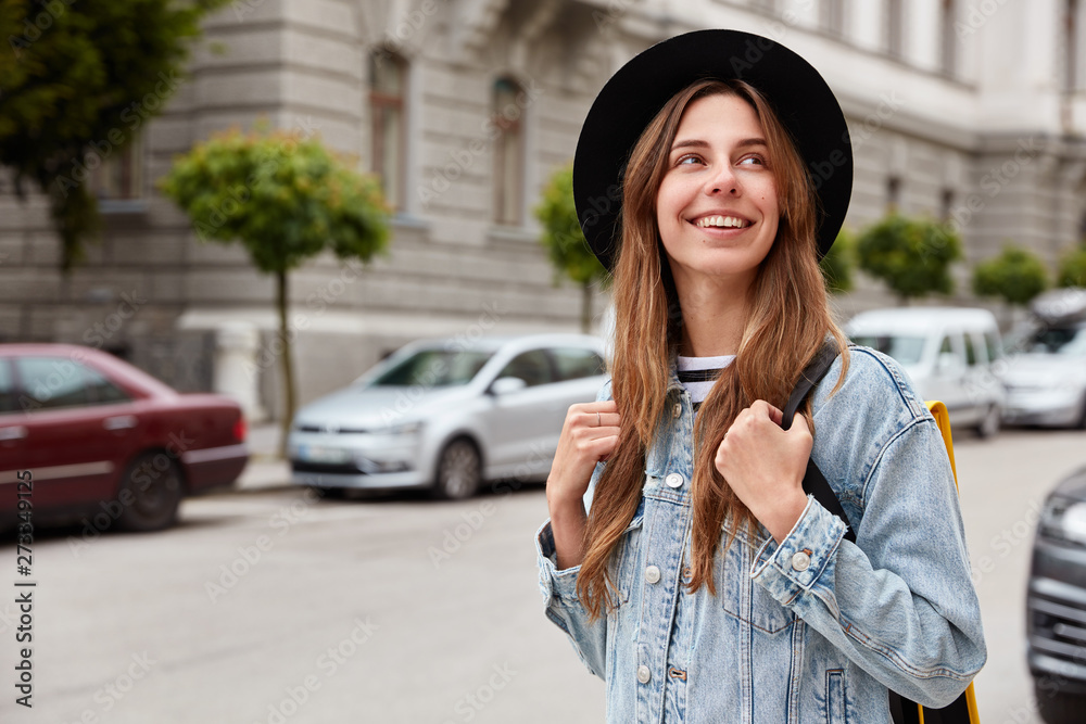Outdoor shot of beautiful European woman strolls through city, spends free time, recreats during vacation, wears hat and denim jacket, walks around cozy street, blurred background, transport.