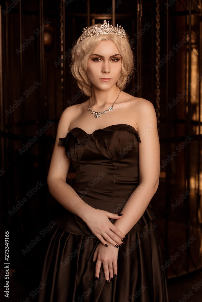 A beautiful, sexy young woman in a black evening dress, with a crown on her head, against the backdrop of a forged cage.