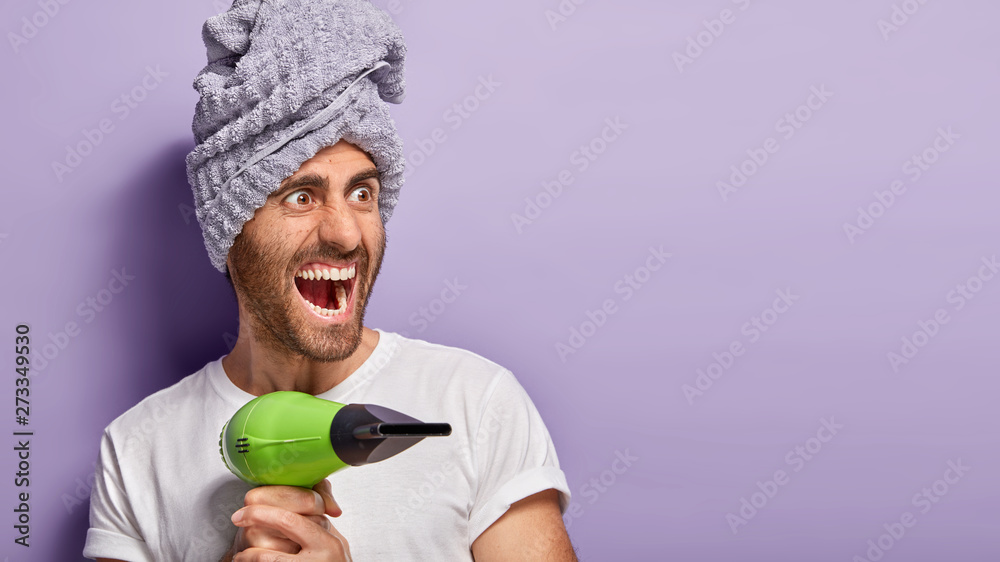 Indoor shot of emotional man with stubble, holds hair dryer, blows hot