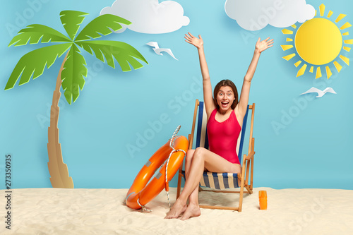 Leinwand Poster Beautiful slim woman raises hands, sits at deck chair on beach, wears red swimwe