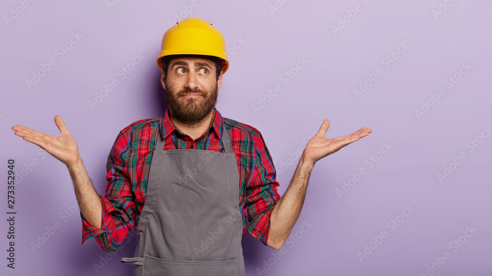 Unaware male construction worker wears yellow protective helmet, checkered shirt and apron, feels doubt which tool to use. Workman or engineer in special uniform being on work, poses indoor.