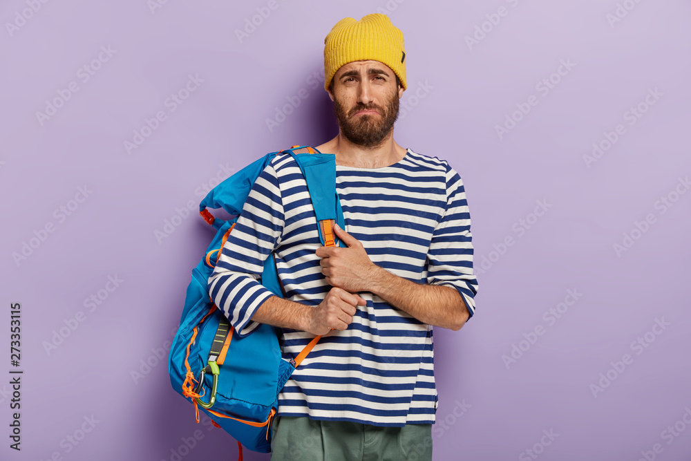 Upset dejected hitchhiker poses with big rucksack, dressed in striped sailor jumper, tired after long journey, looks unwillingly at camera, isolated over purple background. Tourism, adventure concept Upset dejected hitchhiker poses with big rucksack, dressed in striped sailor jumper, tired after long journey, looks unwillingly at camera, isolated over purple background. Tourism, adventure concept