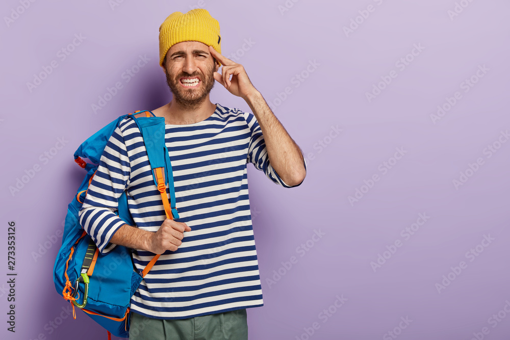Stressful male adventurer has headache after tiring trip, clenches teeth from pain, wears stylish outfit, poses with backpack, cannot concentrate, poses over purple wall, free space for your advert